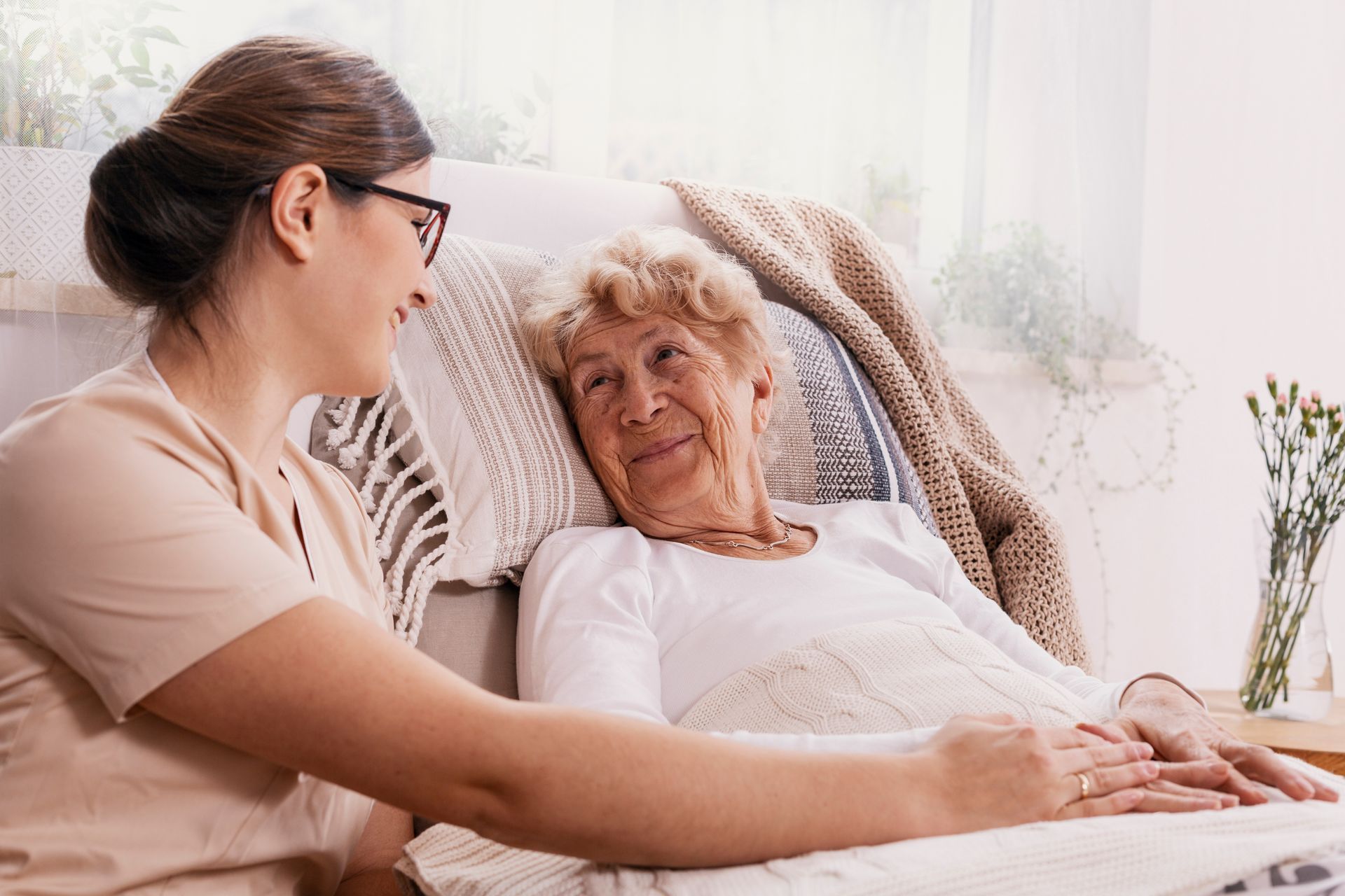 Caregiver smiling at older person in bed; hands touching.