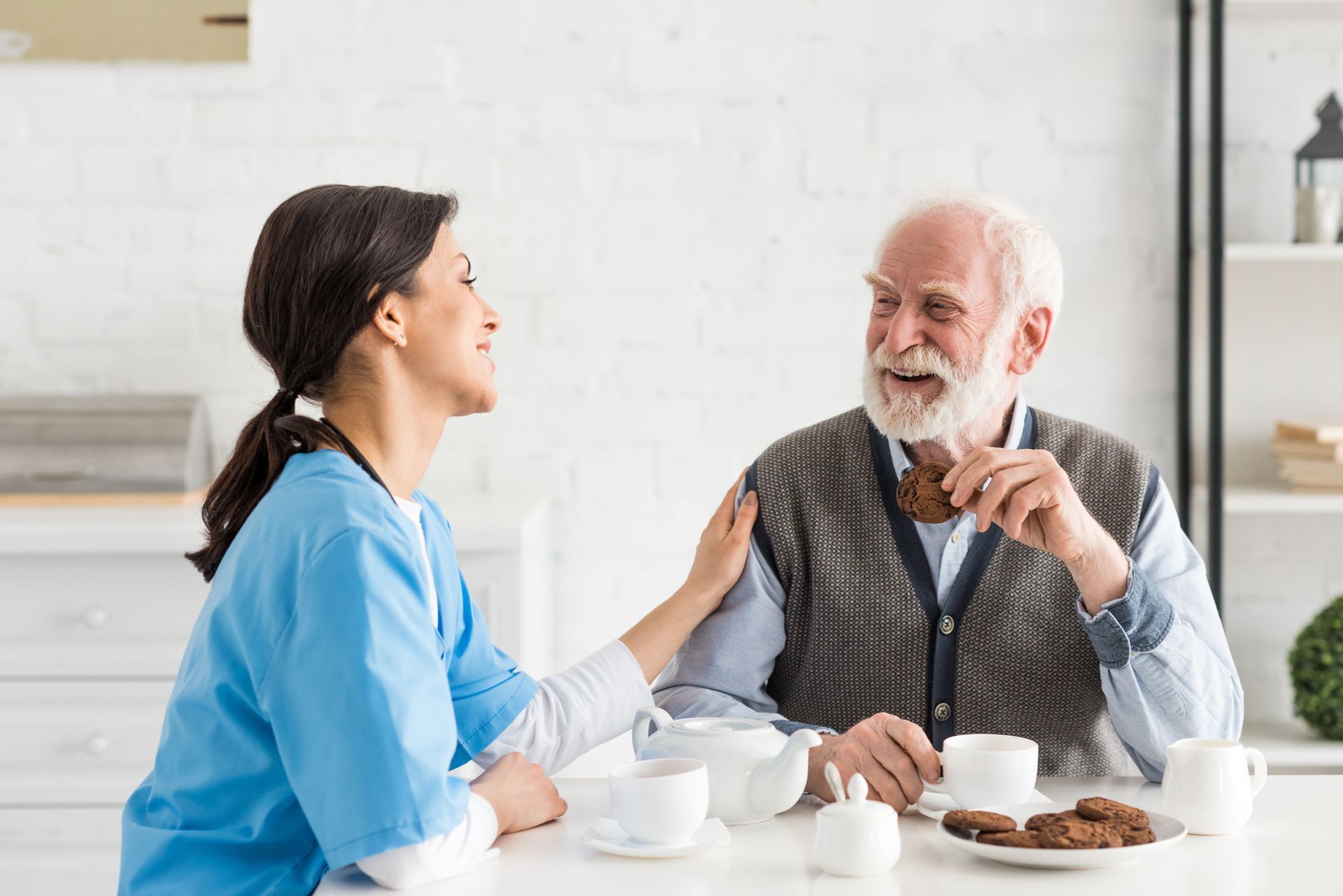 Caregiver with arm on older person's shoulder, smiling, eating a cookie at a white table.