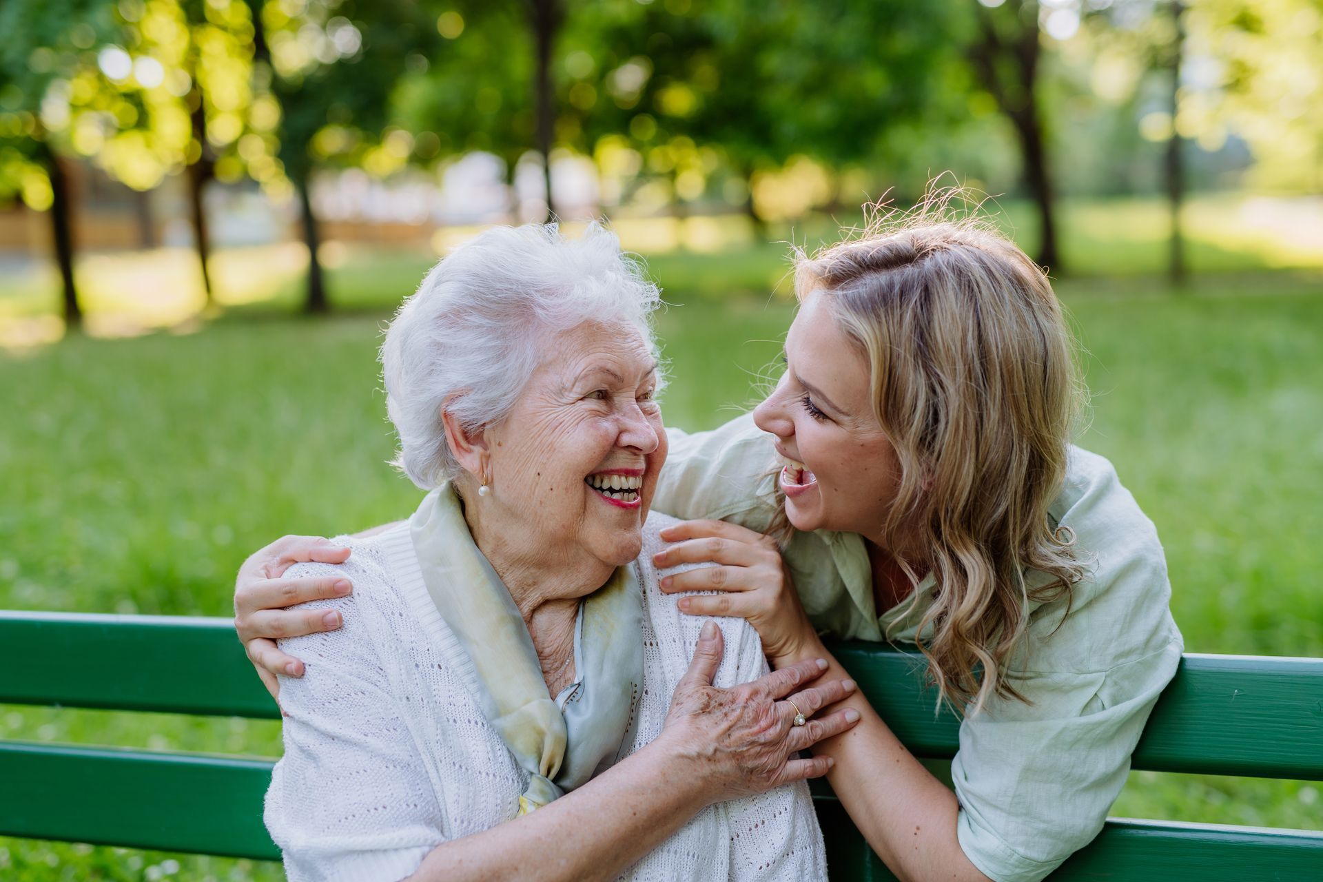 Woman embraces and laughs with older person on a park bench. 