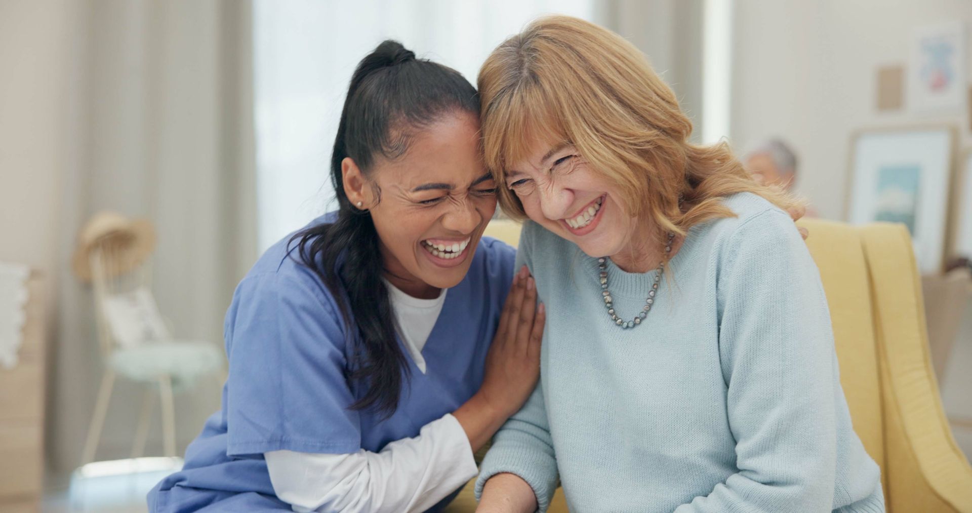 Woman in blue scrubs and older woman laughing together in a brightly lit room.