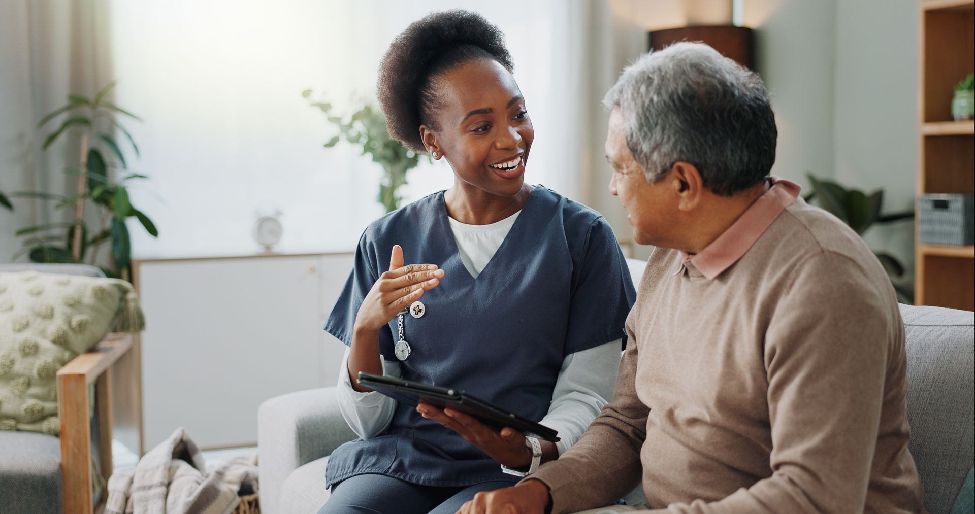 A healthcare worker shows a tablet to a person, both seated on a couch, indoors.
