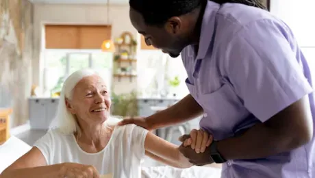 Man in purple shirt assists an older woman, smiling, in a bright room.