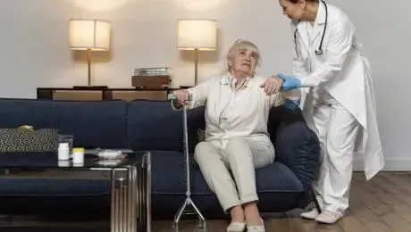 Elderly person with cane helped by healthcare worker in living room.