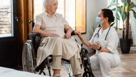 Nurse in mask kneels, touching the arm of a person in a wheelchair, indoors.