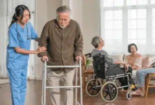 Nurse assisting a man with a walker, another in a wheelchair, and two others sitting in a living room.