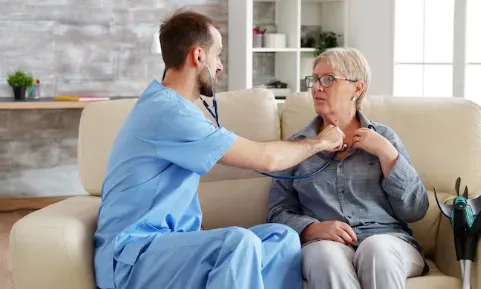 A healthcare worker listens to an older person's chest with a stethoscope while they sit on a couch.