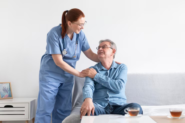 Nurse in blue scrubs comforts older man on a couch, indoors, holding his arm.