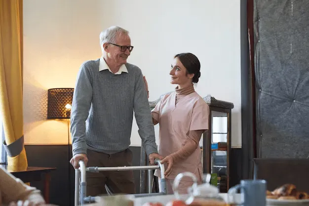 Elderly person using walker, assisted by a healthcare worker indoors, smiling.