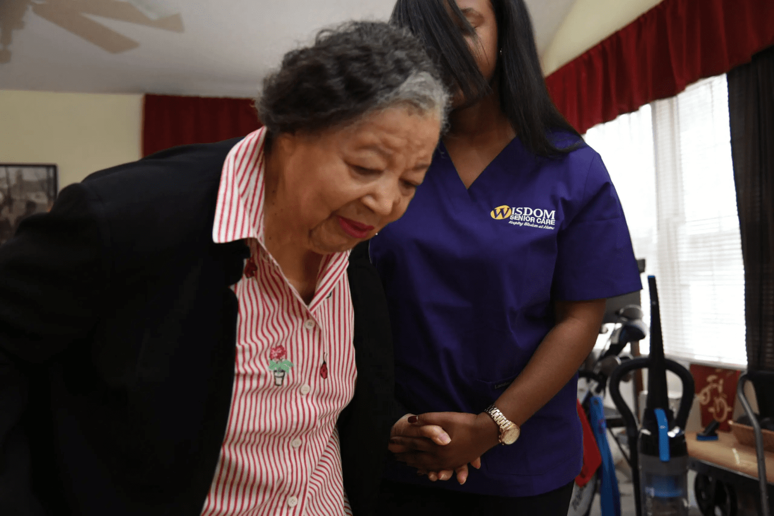 Older person assisted by a person in a purple scrub in a room.