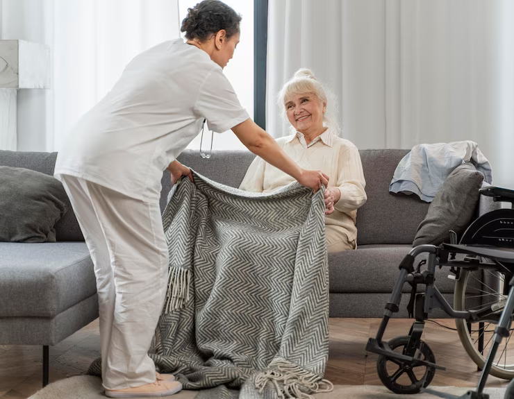 Caregiver placing blanket on woman seated on couch. Indoors with wheelchair.