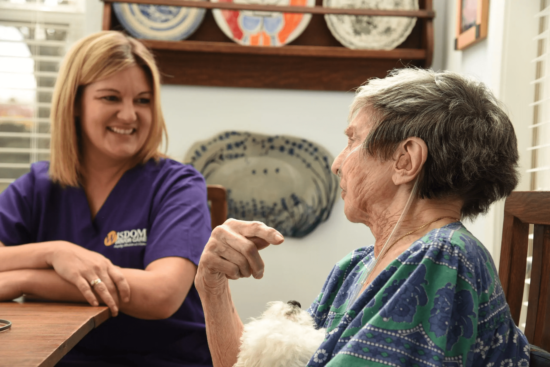 Caregiver smiles at an elderly person seated at a table. 