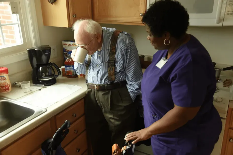 An older person sips from a mug while a caregiver assists in a kitchen.