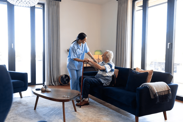 Caregiver assisting an older person to sit up on a blue couch in a living room.
