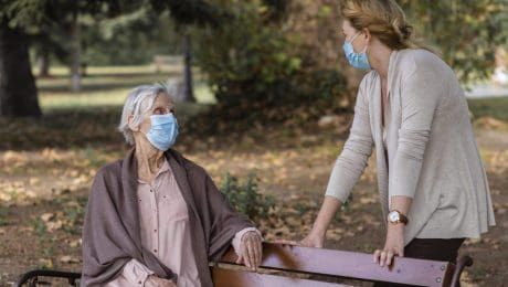 Woman in mask talks to an older person wearing a mask, sitting on a park bench.