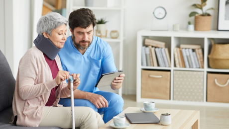 Woman with neck brace and cane consults with healthcare provider, both looking at tablet in living room.