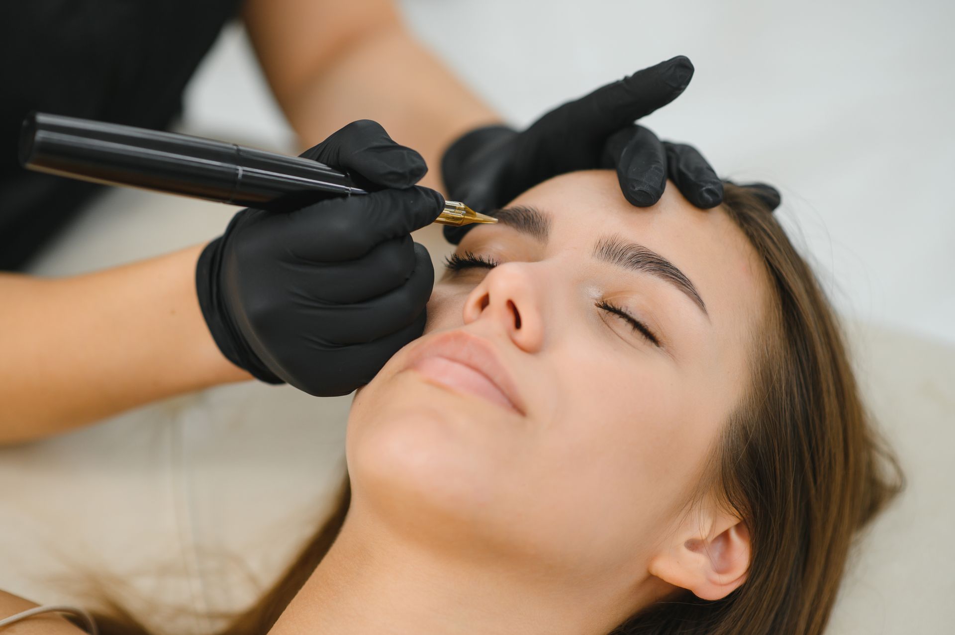 Person receiving an eyebrow microblading treatment; technician using a handheld tool.