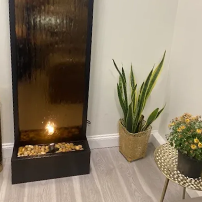 An indoor water fountain next to a potted snake plant and a gold side table with flowers.
