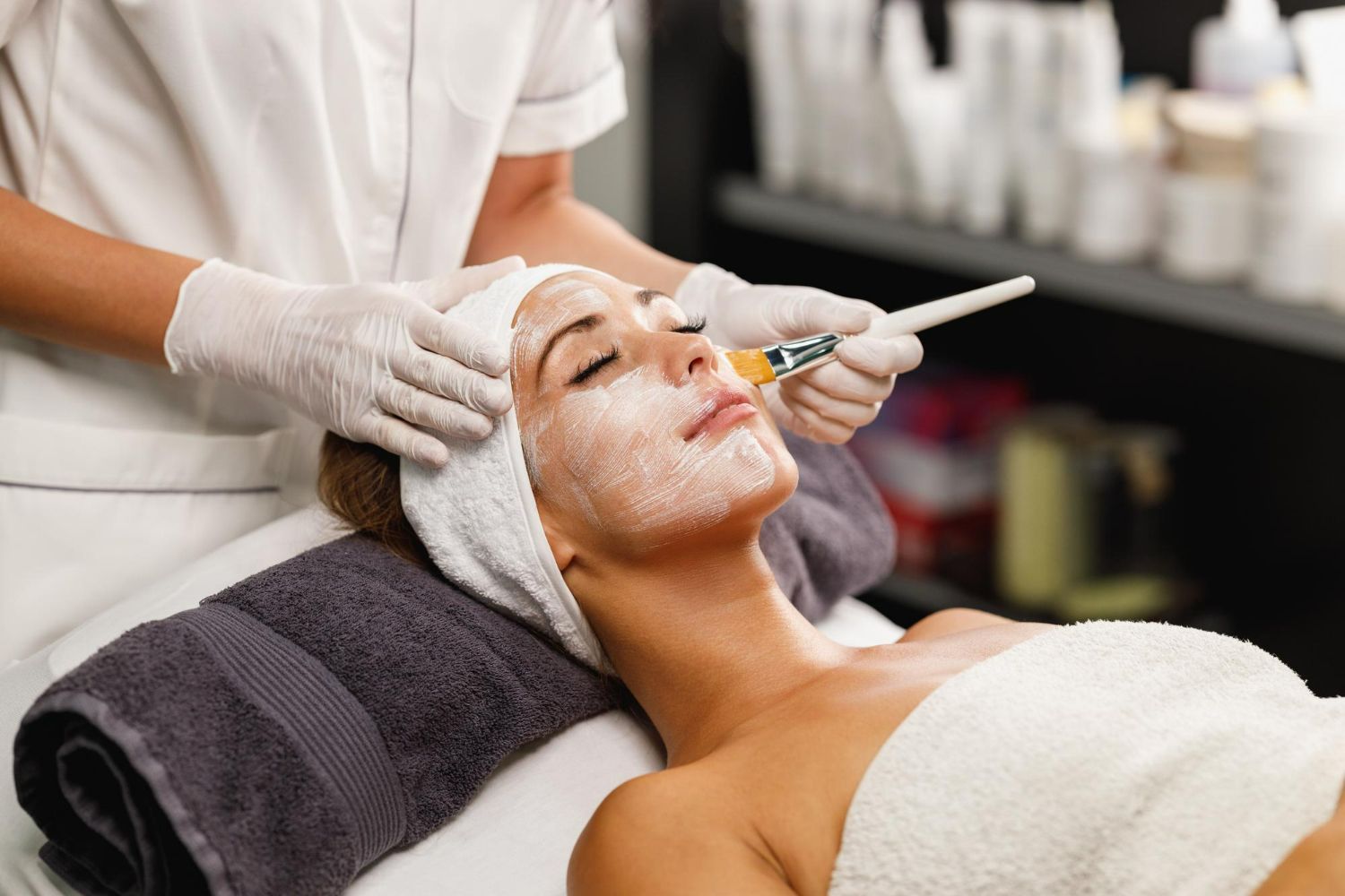 Esthetician applying facial treatment to a client in a spa, with white cream on the face and a towel headband