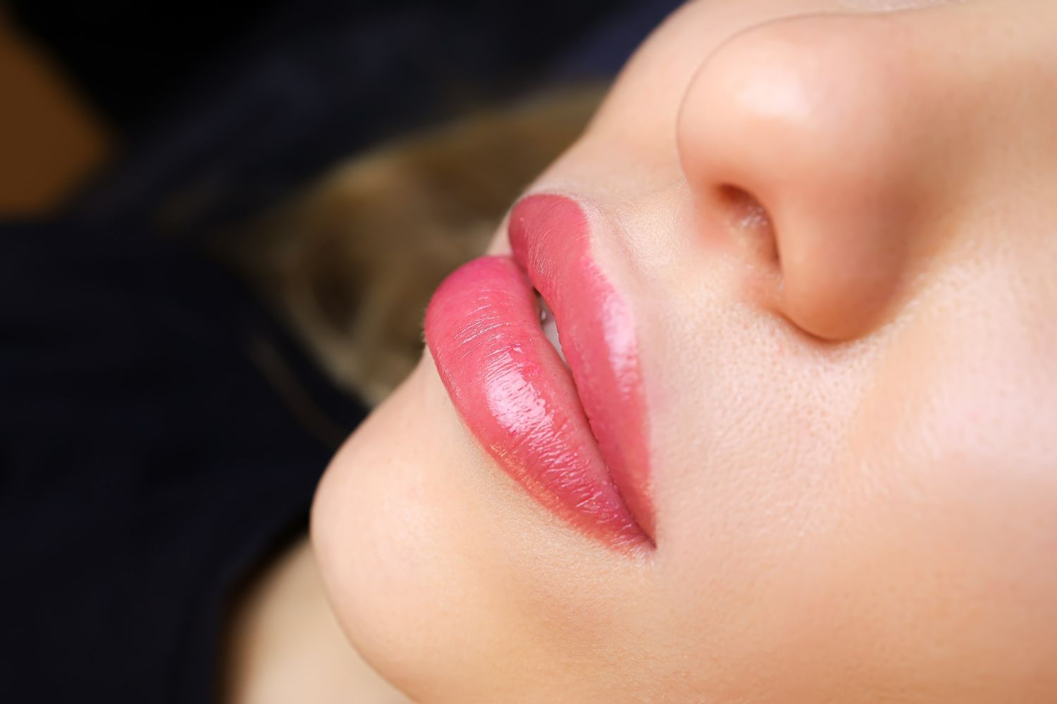 Close-up of glossy pink lips and part of a nose against a dark background