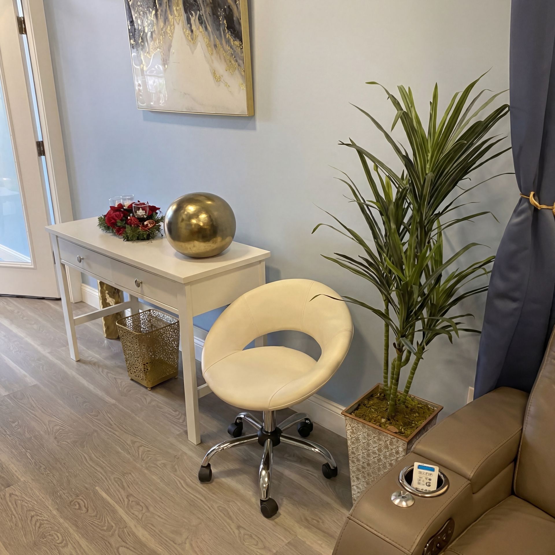 A white console table with a gold sphere, a small chair, a tall potted plant, and part of a tan recliner in a room.