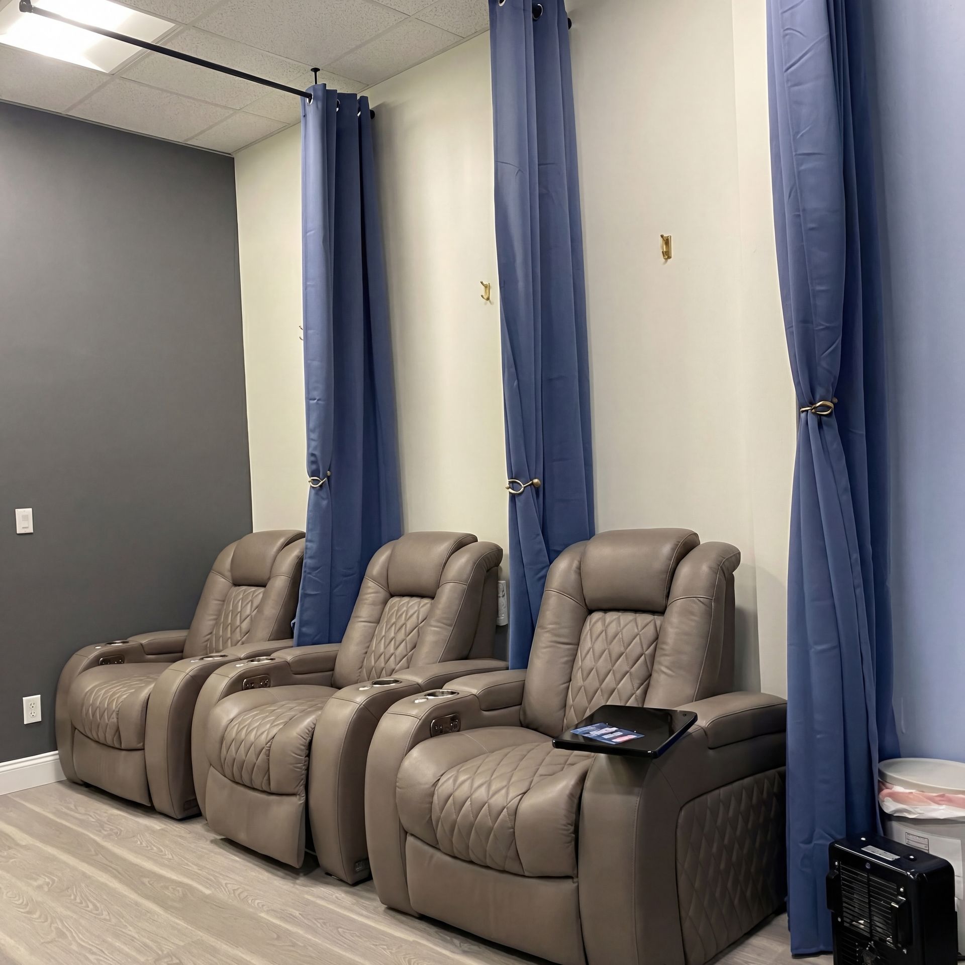 Three taupe leather reclining chairs sit in a row against a wall with blue curtains and light wood-look flooring.