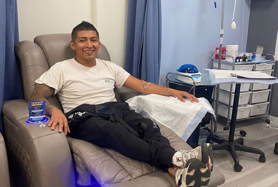 A person smiling while sitting in a clinical recliner, with medical equipment and a water glass nearby.