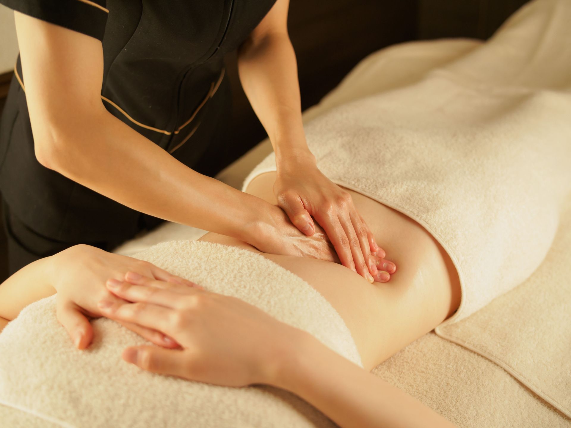 Hands giving a massage to a person's lower back, covered with a white towel, spa setting.