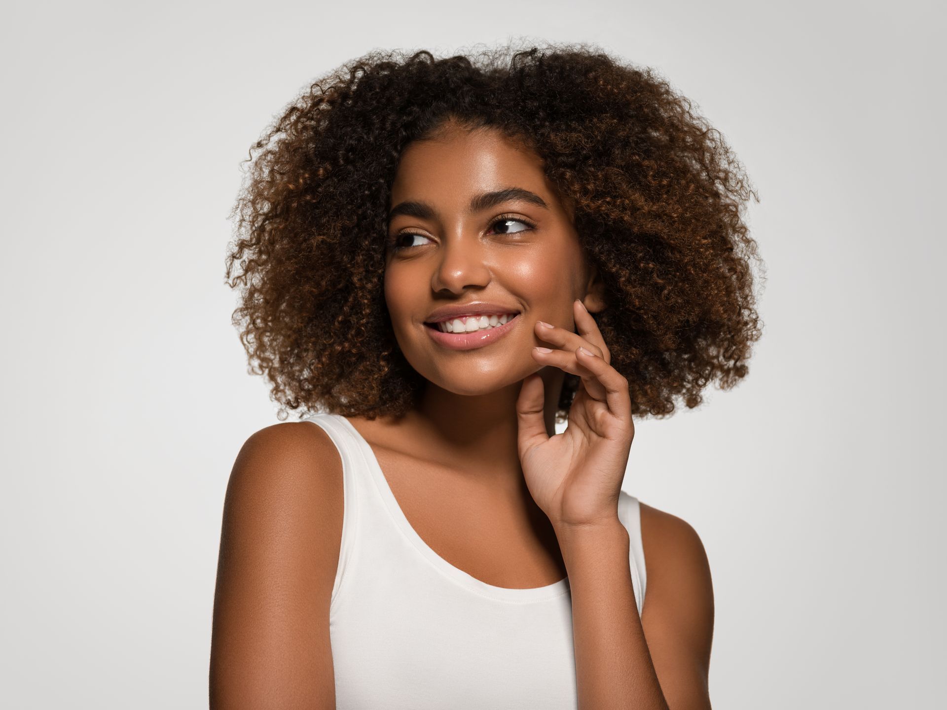 A woman with curly hair is smiling and touching her face.