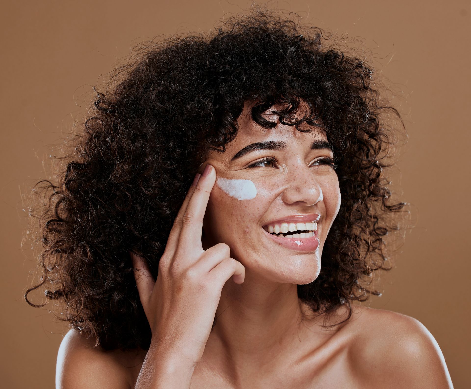 A woman with curly hair is smiling while applying cream to her face.