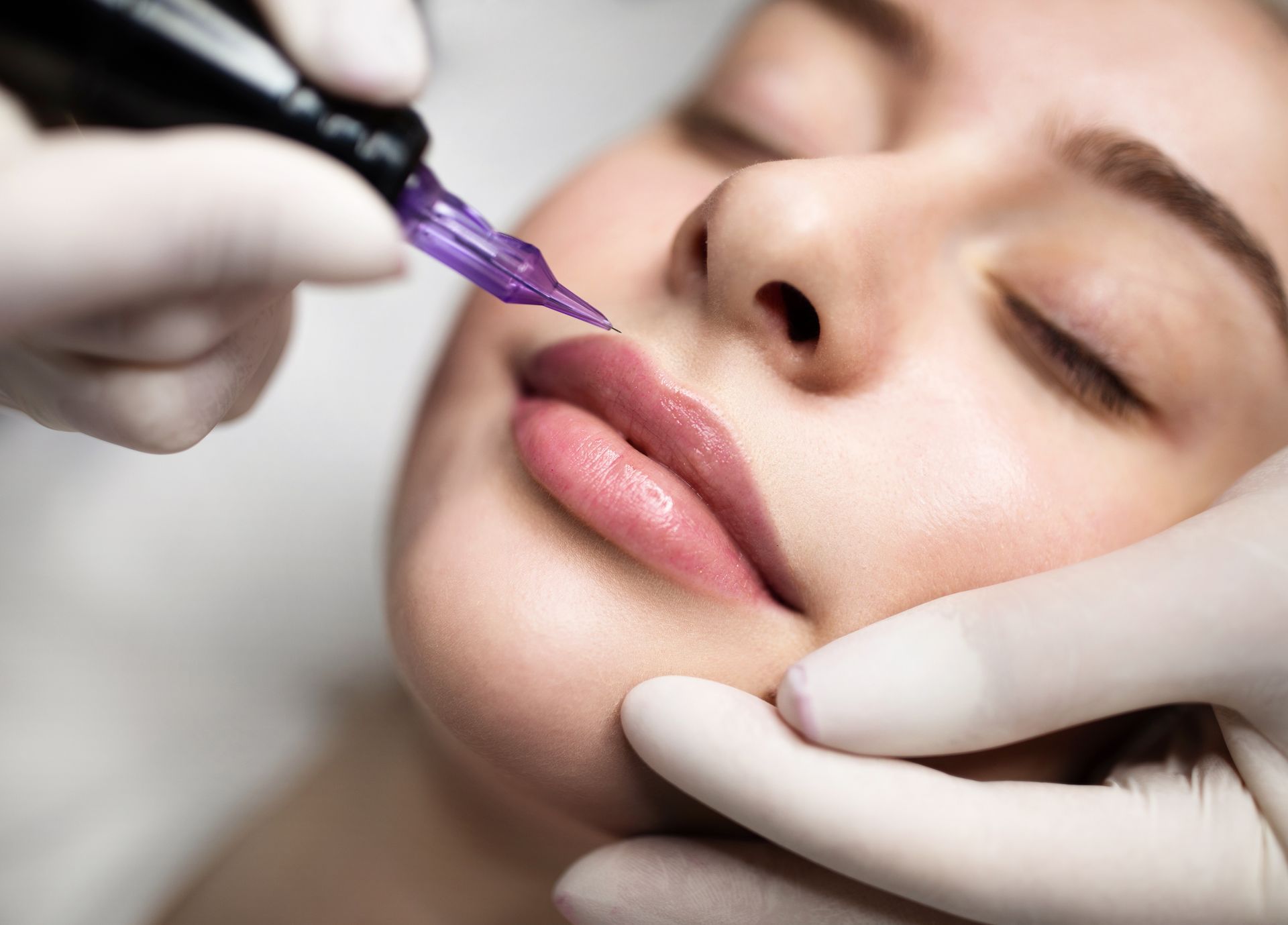 Lips being tattooed by gloved hands, close-up. Light pink lip color with closed eyes.