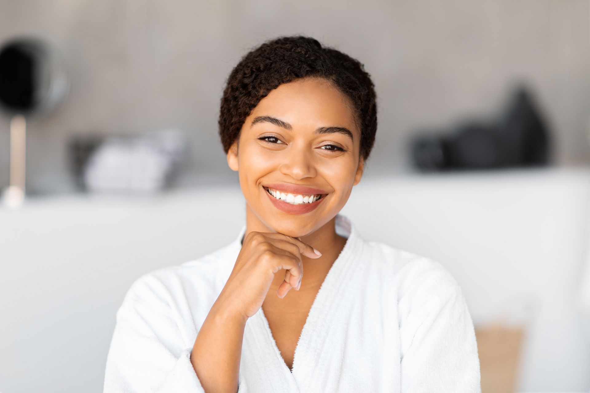 Woman in white robe smiling, resting chin on hand. Blurred bathroom background.