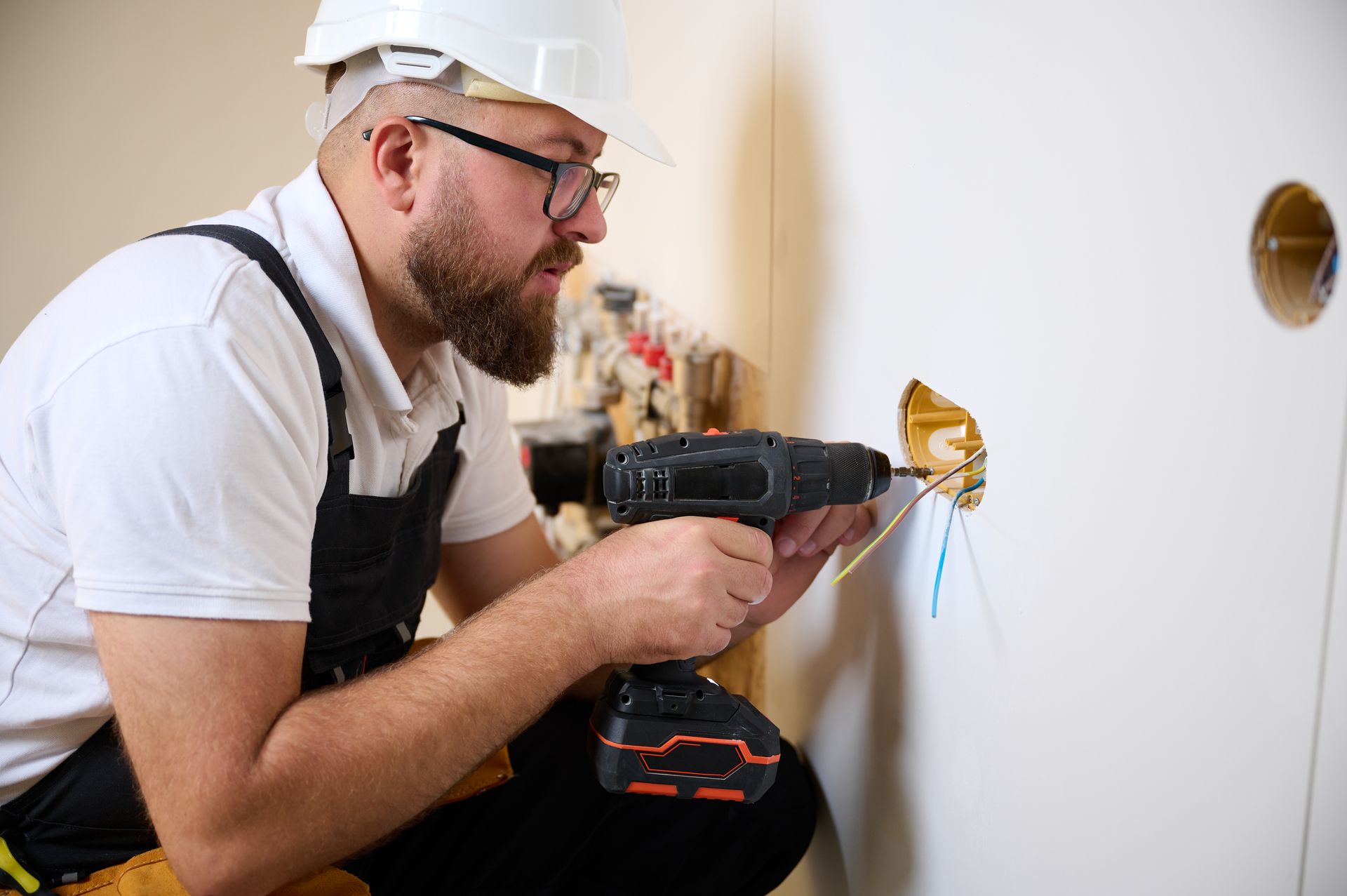 Electrician in a white hard hat installs an outlet using a drill on a white wall.