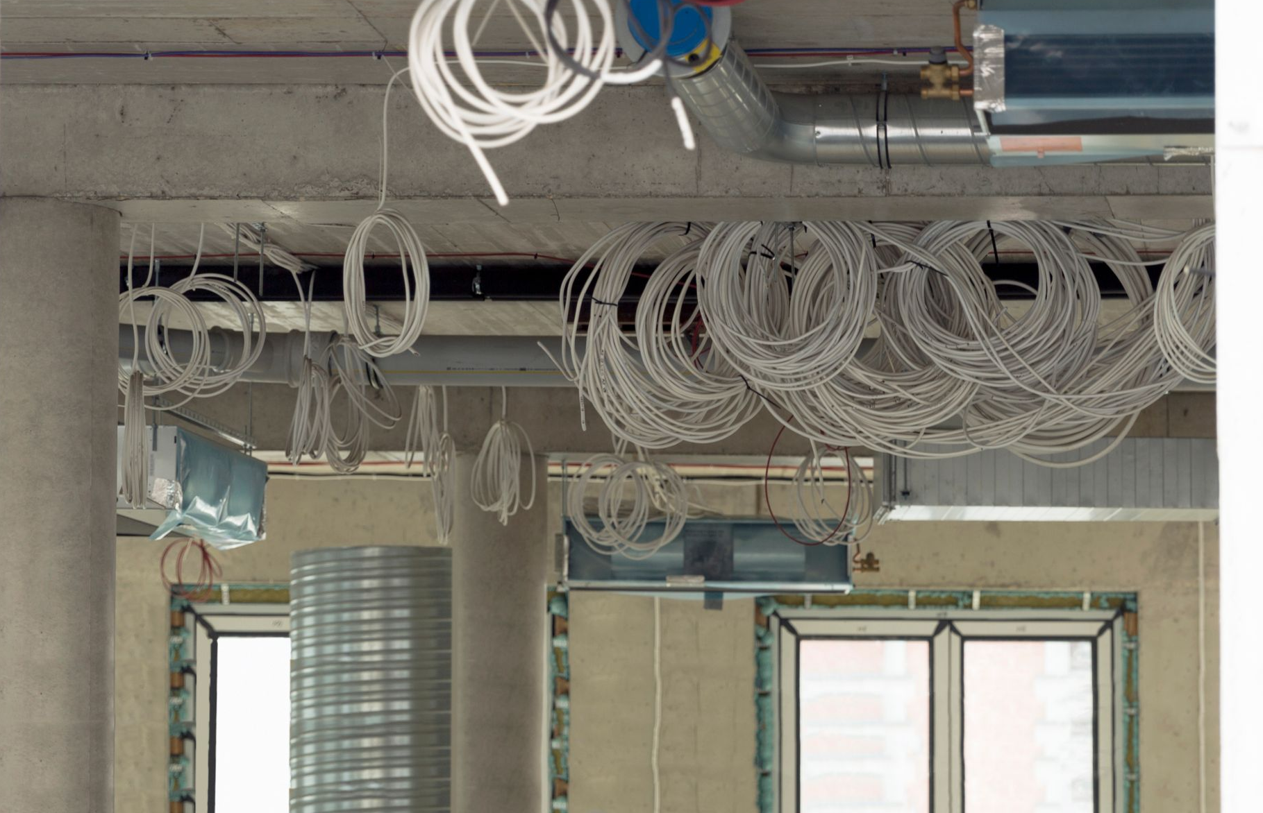 Construction site interior with exposed wiring, ductwork, and concrete ceiling near windows.