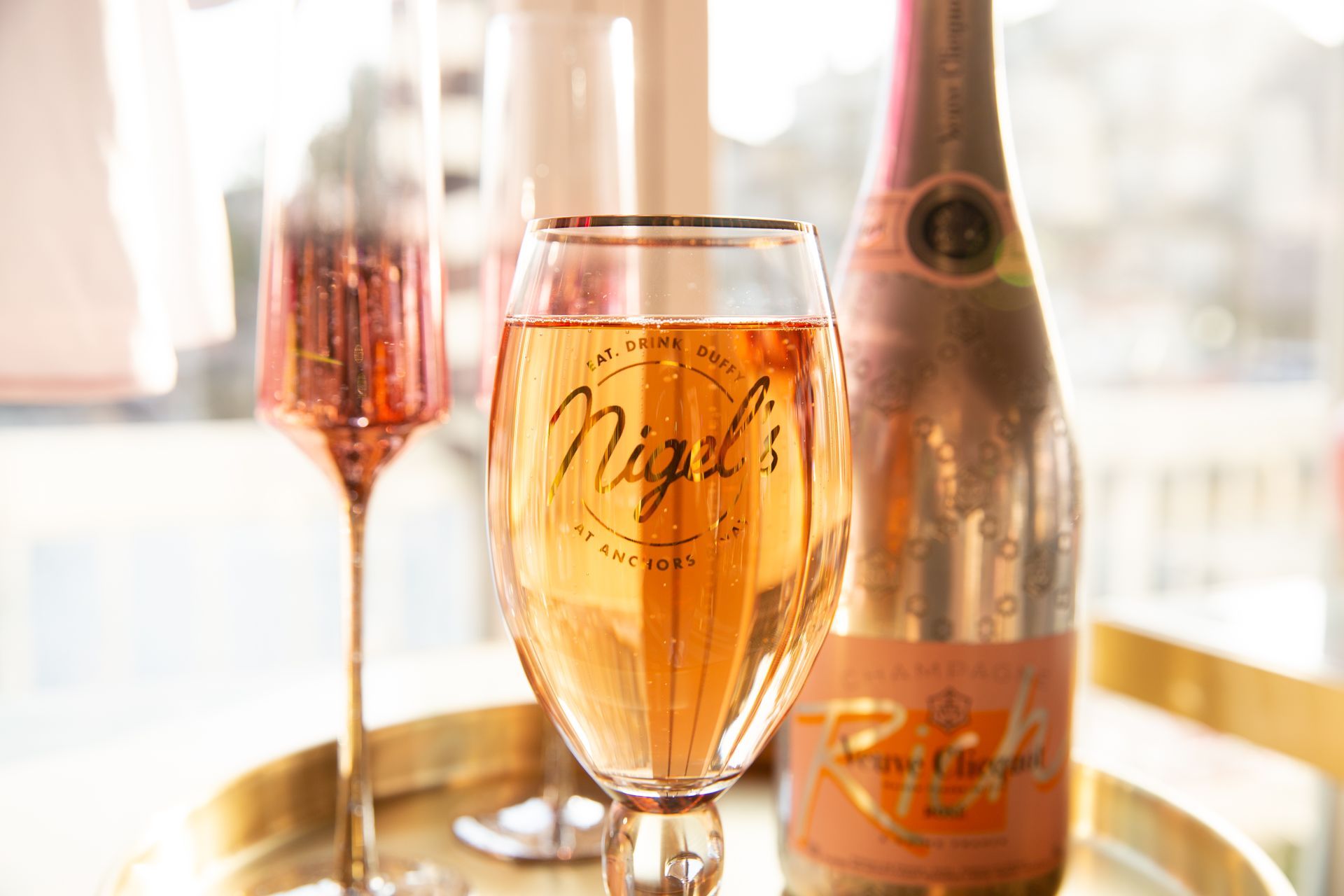 Three champagne flutes with rosé on a sunlit table beside a bottle of sparkling wine.