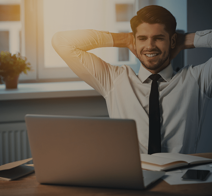 Um homem está sentado em uma mesa com as mãos atrás da cabeça em frente a um laptop.