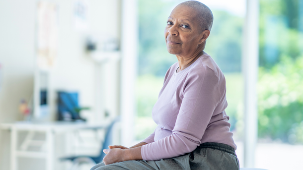 Woman with shaved head in a pink shirt sits in a light-filled room, smiling slightly.