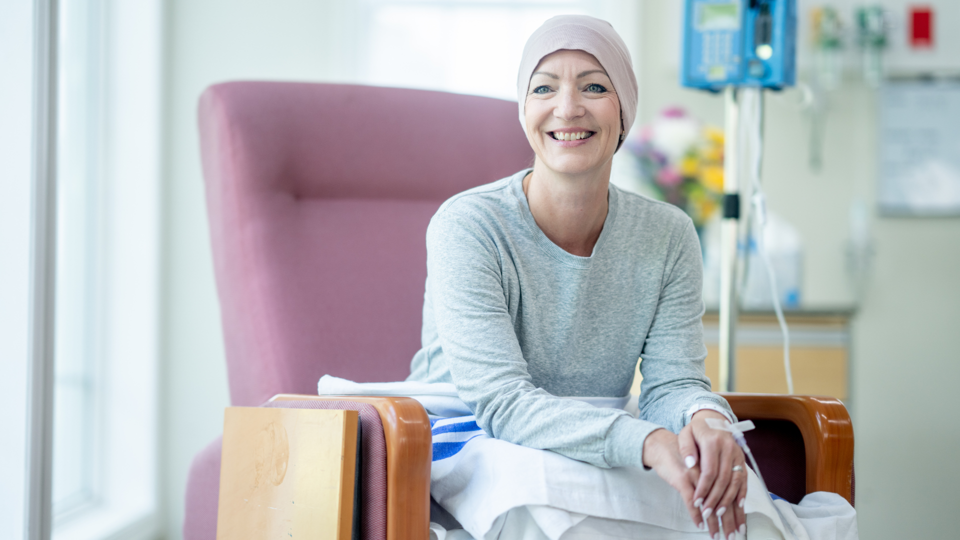 Woman in headscarf smiles, sitting in a pink chair in a hospital room.