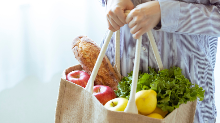 Person holding a reusable bag filled with groceries: apples, lettuce, and bread.