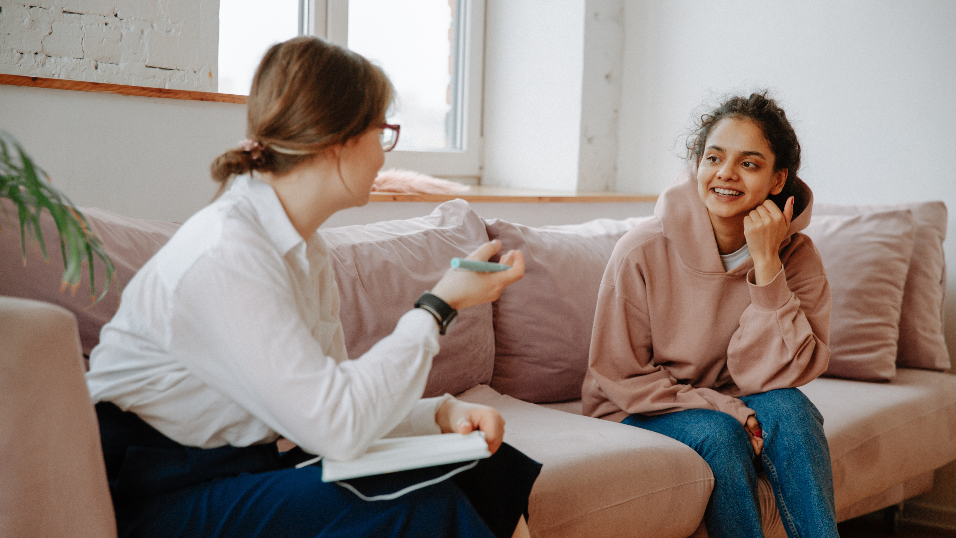 Woman on couch smiling, talking with another woman holding a notebook.