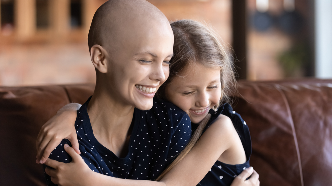 Woman with bald head smiles, hugging a young girl on a brown couch.