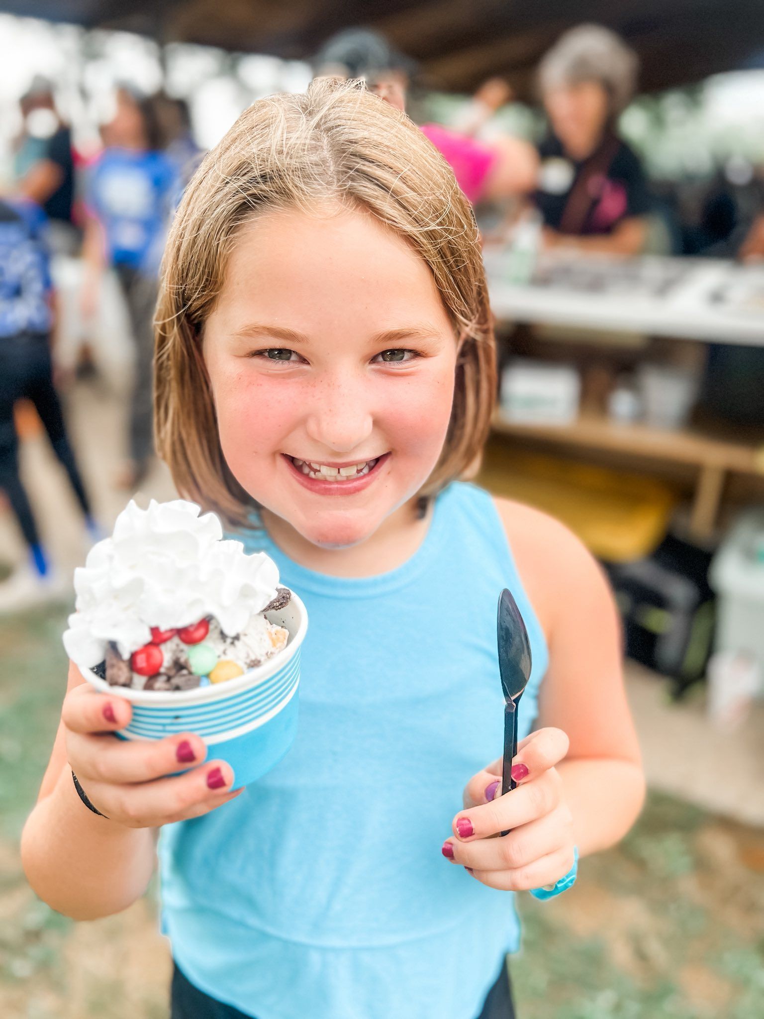 Girl with a big smile, holding an ice cream sundae topped with whipped cream and a small black spoon.