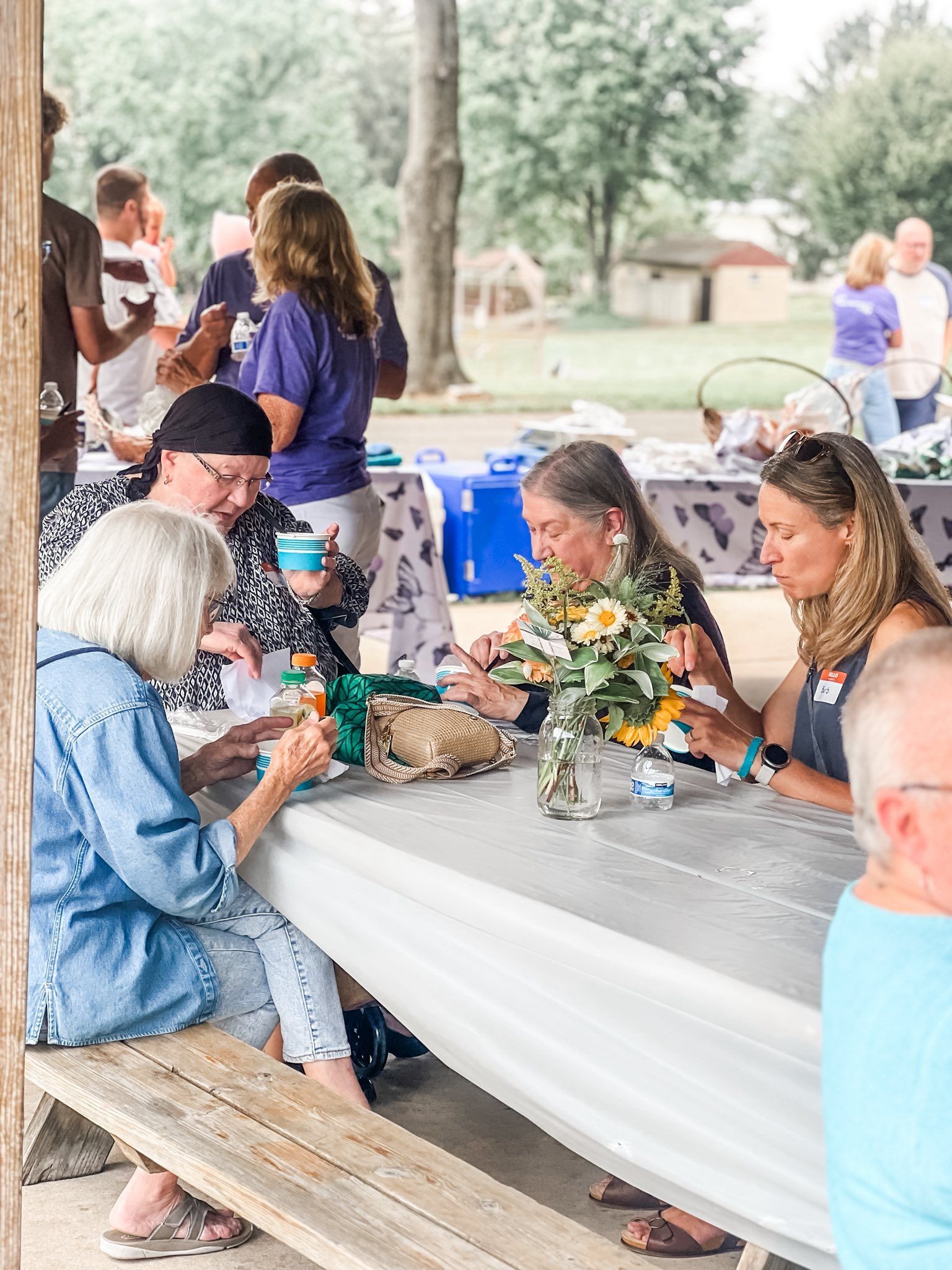 People at a picnic: seated at a table, some eating and drinking, others in the background. Outdoor setting.