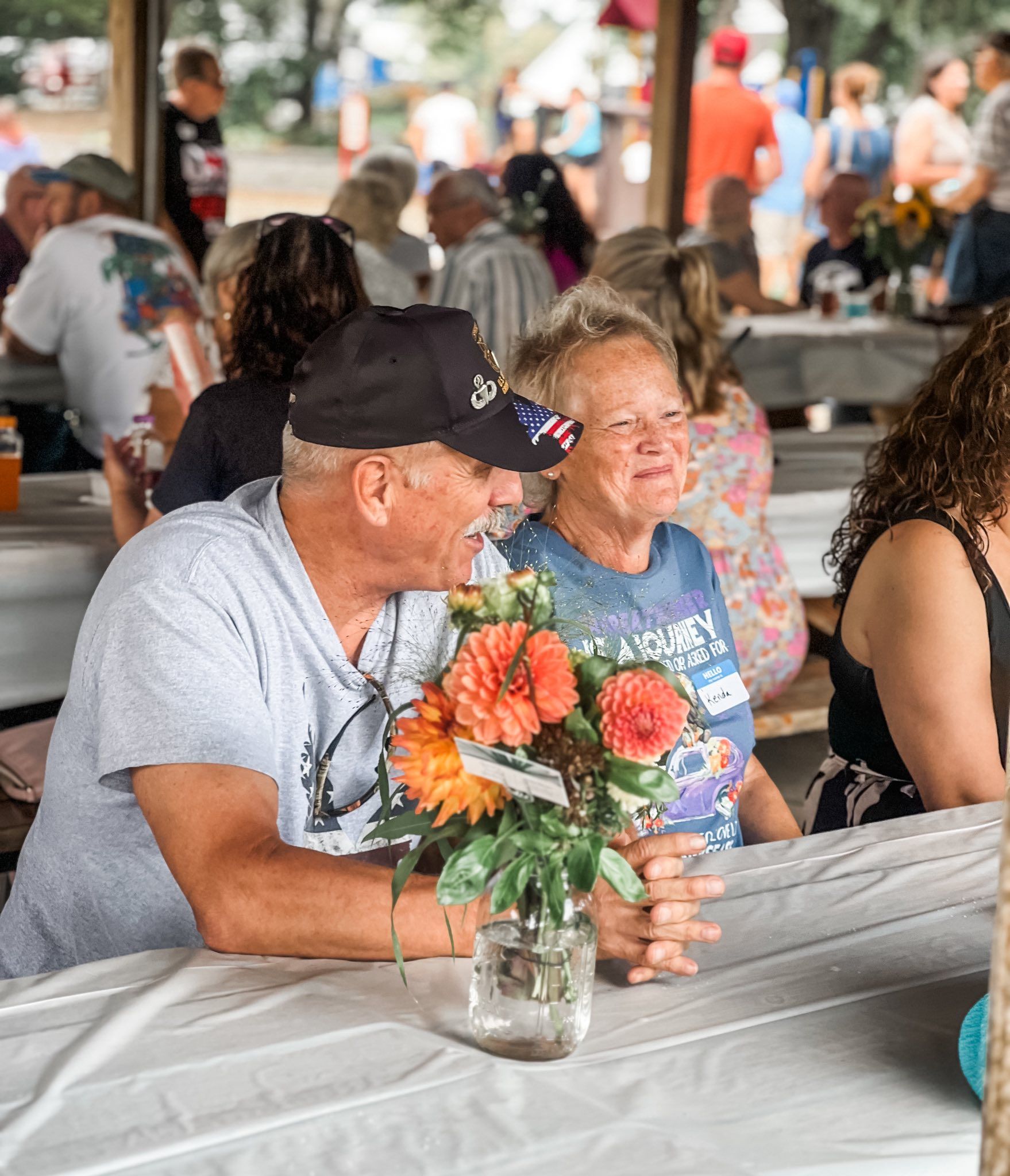 Couple at a picnic table, smiling. Flowers in a jar. Other people in the background.