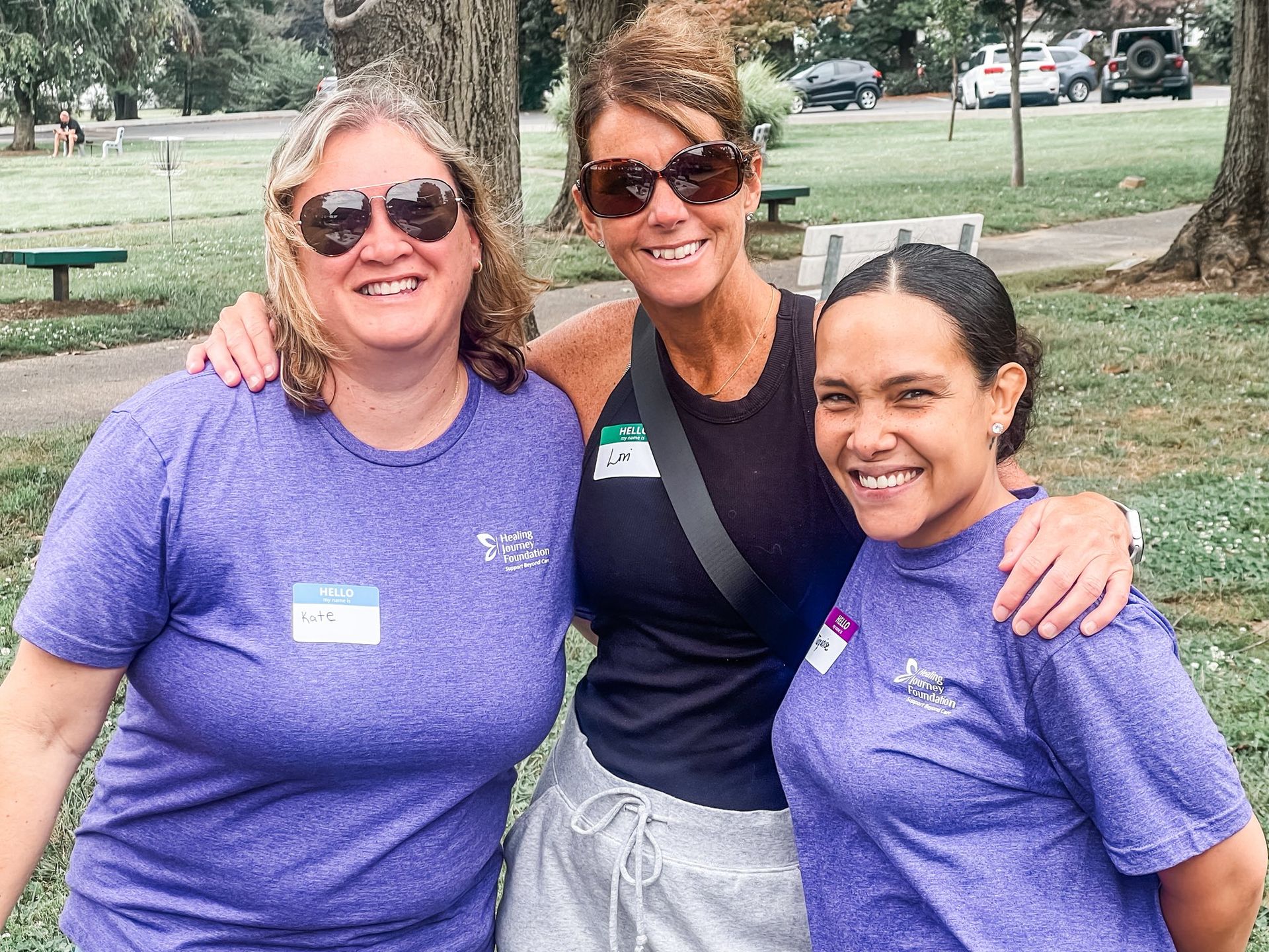 Three people smile, arms around each other. Two wear purple shirts, one black tank top, outdoors.