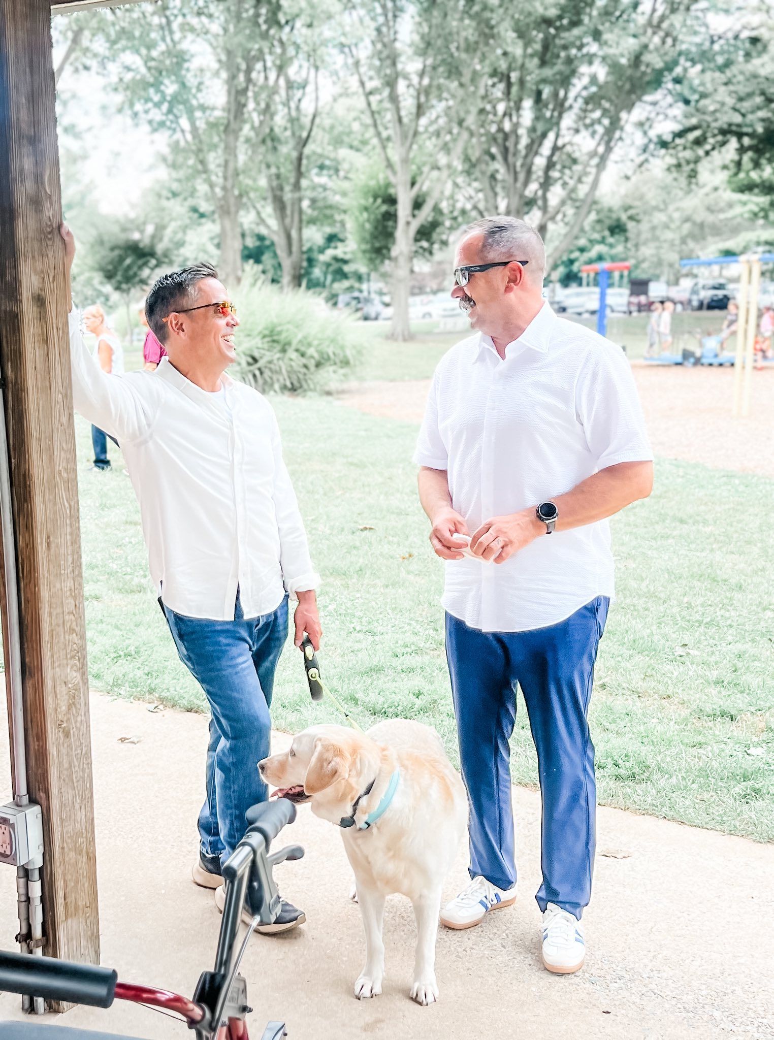 Two men in white shirts and jeans talking, with a golden retriever dog, in a park setting.
