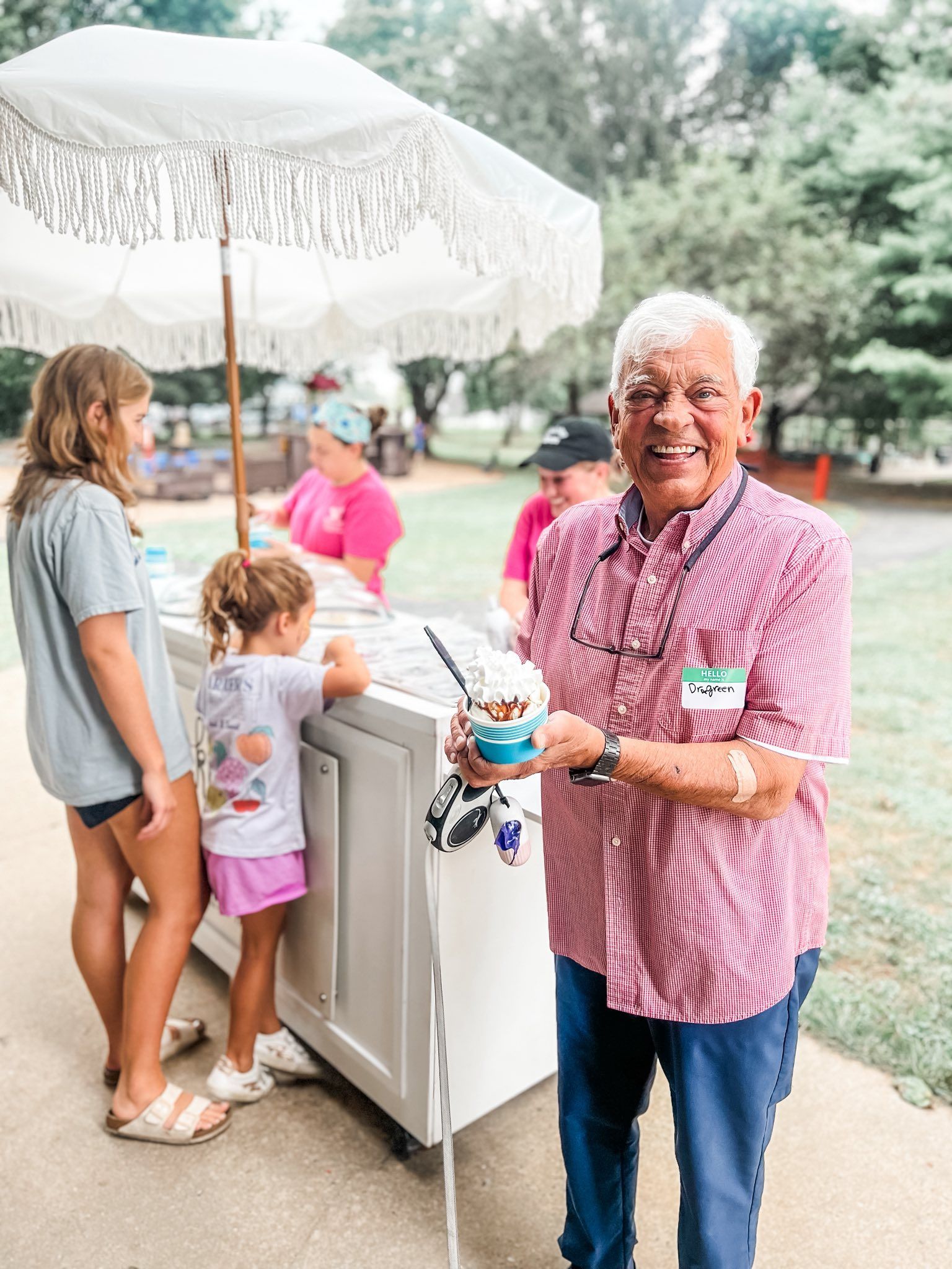 Man holding ice cream, smiling near an ice cream stand, with two children. Outdoor setting.
