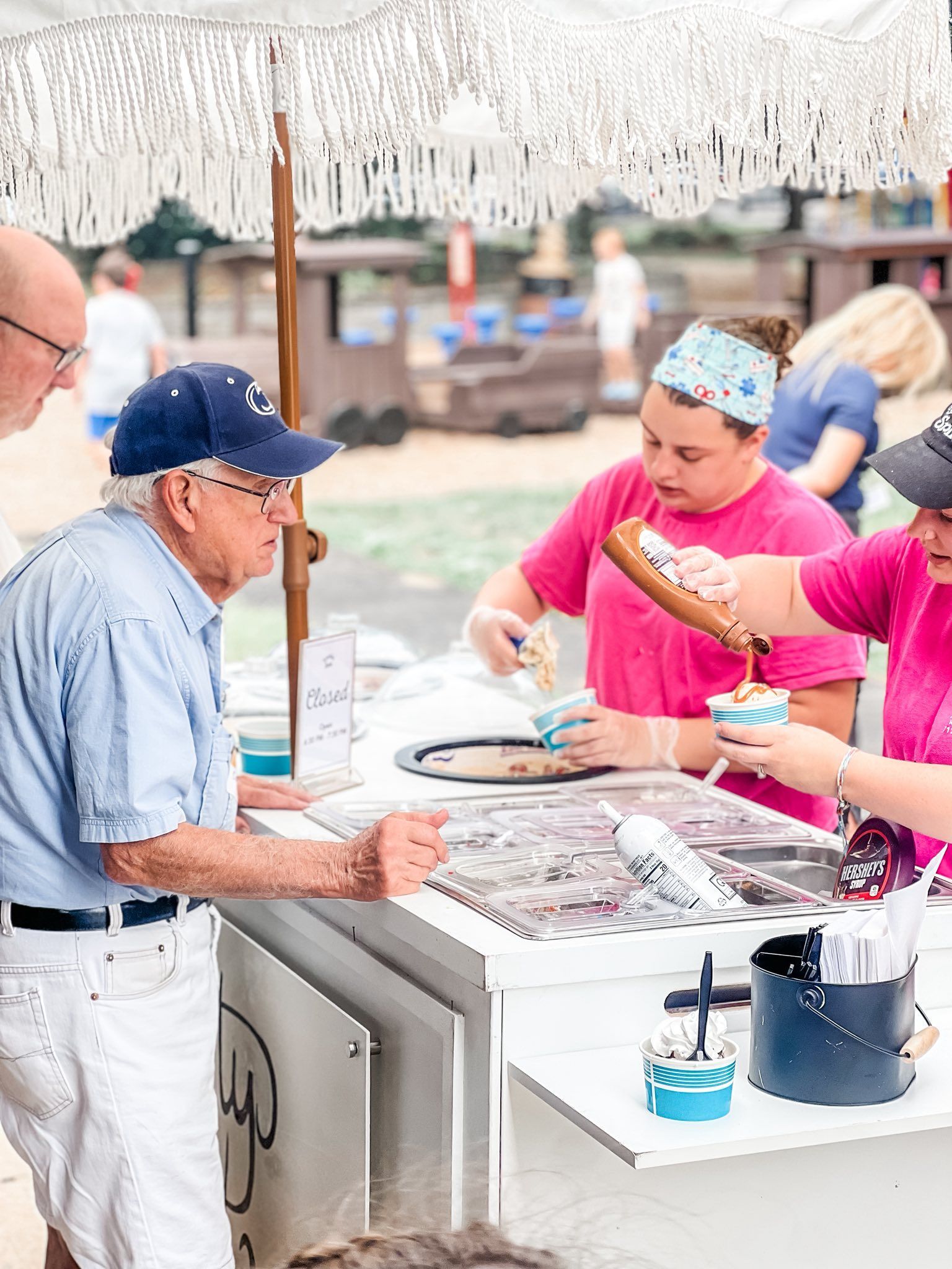 People ordering ice cream from a stand. Two people in pink shirts serve a customer in blue. White parasol overhead.