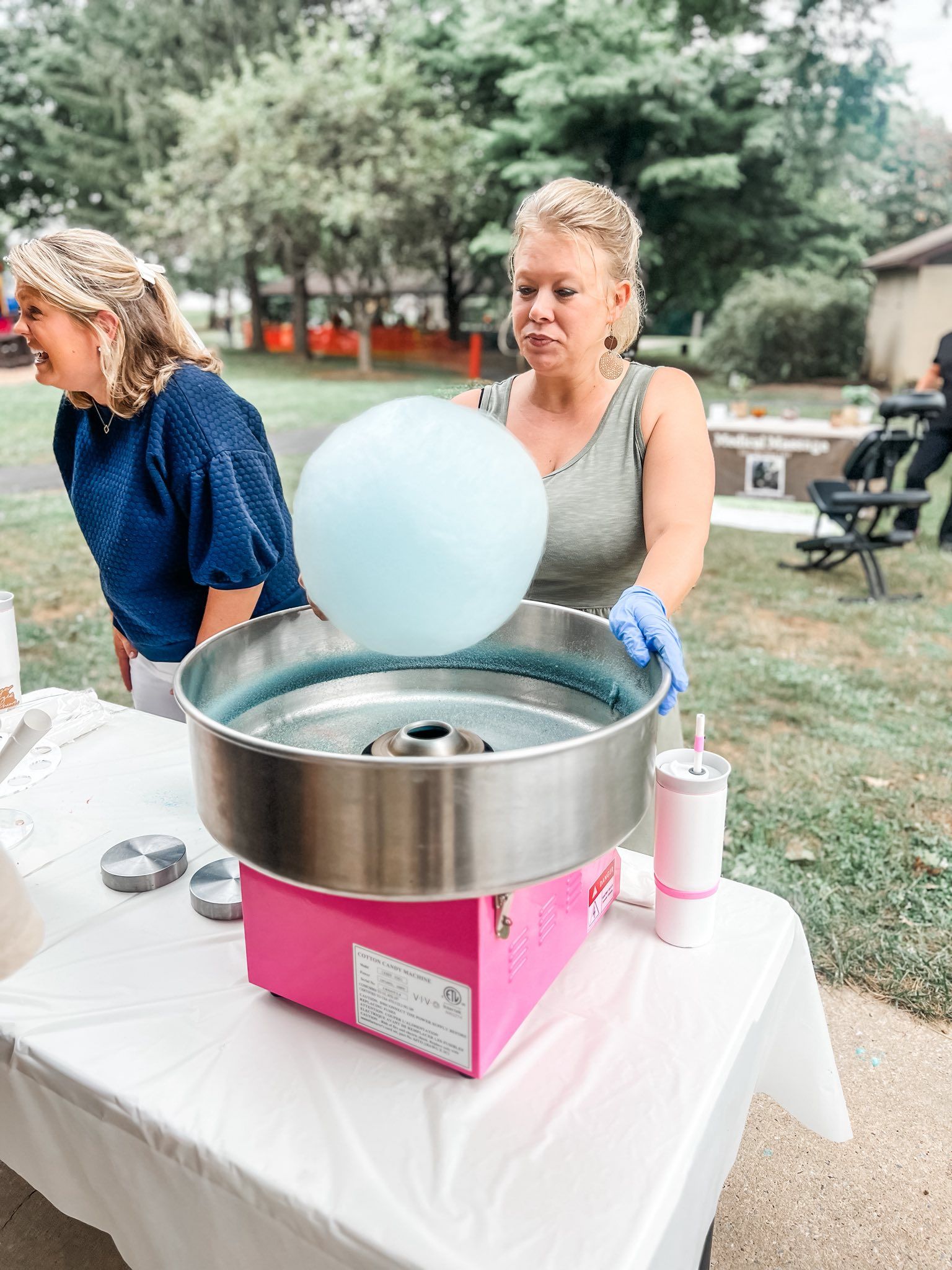 Woman making blue cotton candy at an outdoor event, pink machine, second person smiling nearby.