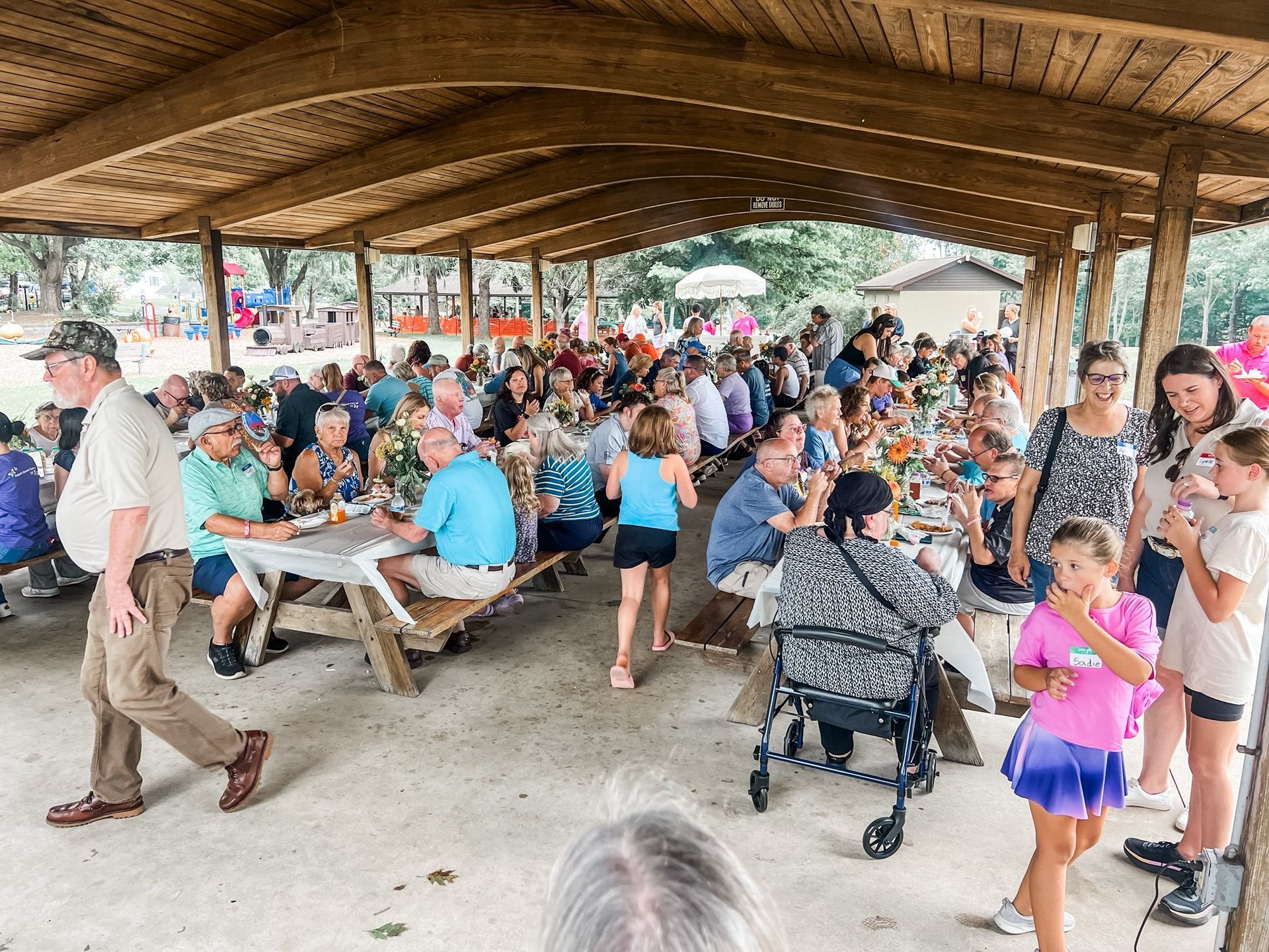Picnic under a wooden shelter. Many people seated at tables, eating and talking. Children running around.