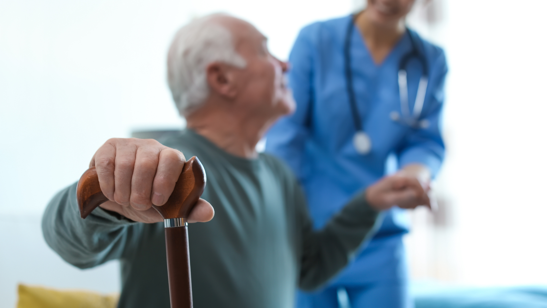 Senior man holding cane, looking toward a healthcare worker in blue scrubs, indoors.